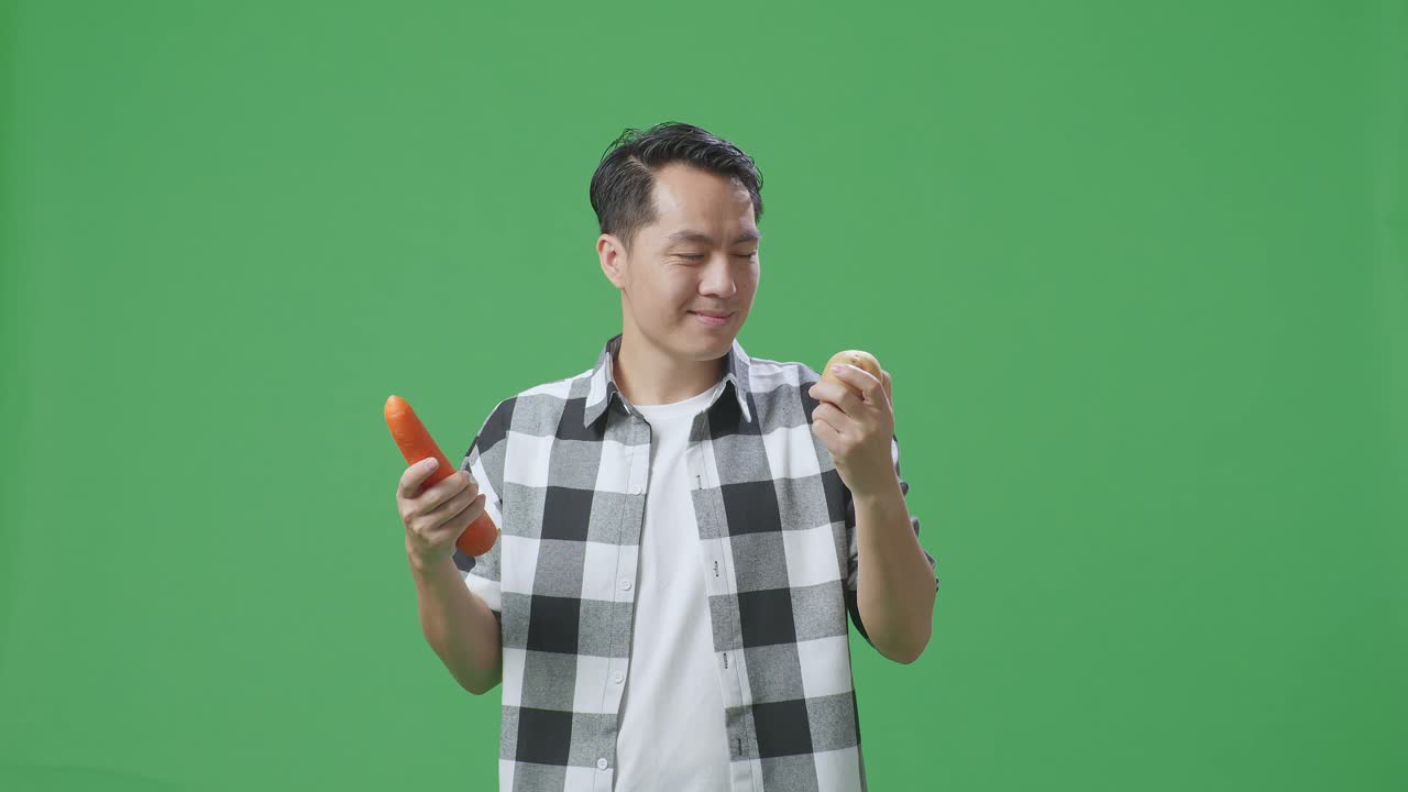아시아 남성 농부 는 스튜디오 의 초록색 스크린 배경 에 따뜻 한 미소 를 지으면서 감자 와 당근 을 들고 있다