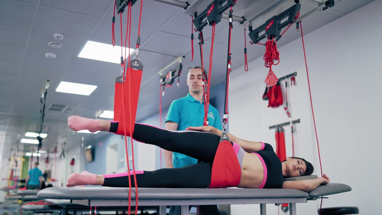 Personal trainer is helping his female client at the modern rehabilitation center. Young woman is lying on a special table on one side and doing exercises with suspension ropes.