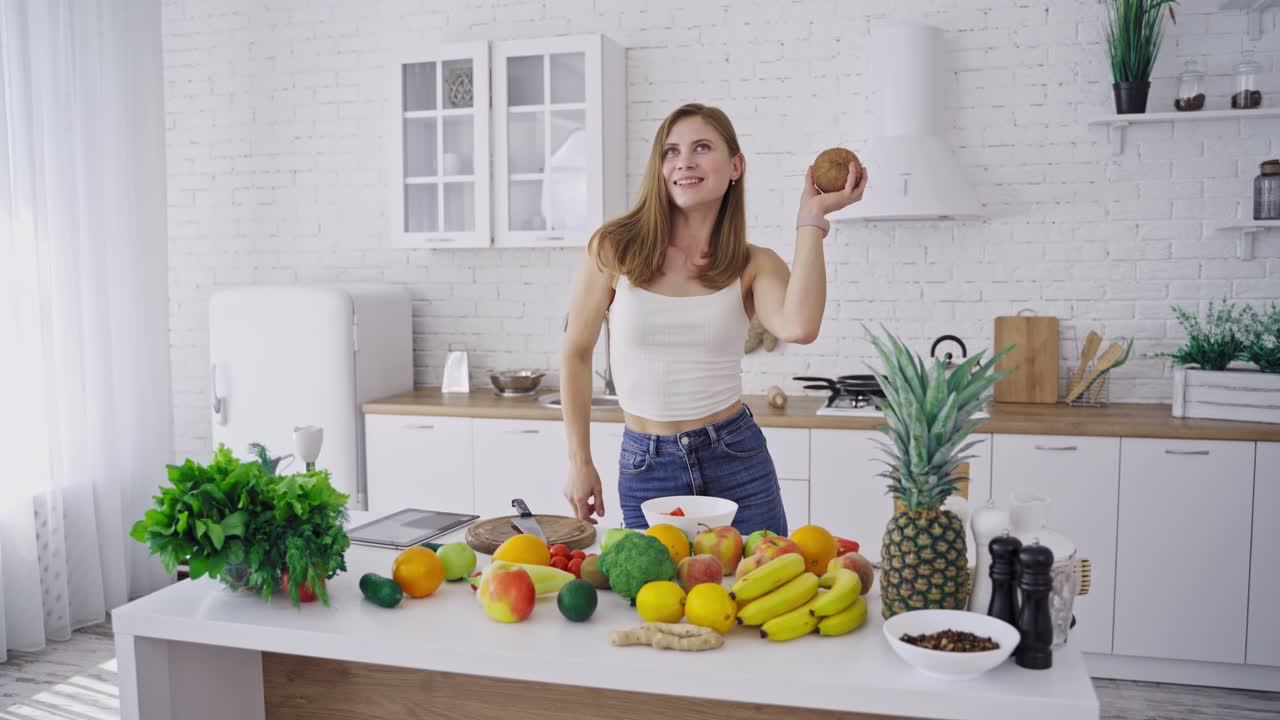 Joyful woman with coconut in the kitchen. Attractive female is dancing with fresh coconut on the background of organic fruit and vegetables. Healthy lifestyle.