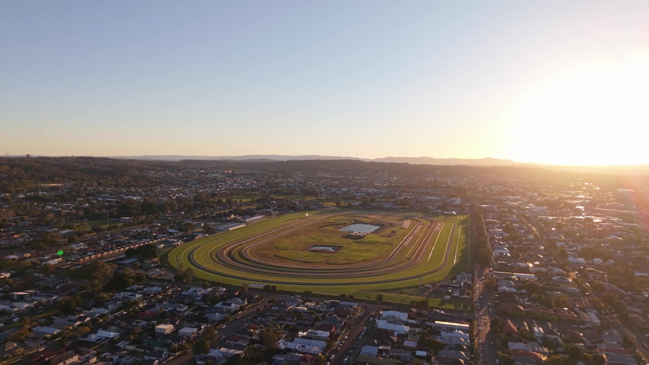 Newcastle Væddeløbsbane og omkringliggende forstæder gløder i lavt aftensolskin ved solnedgang, et panoramisk luftfoto med tilbagetrækning i den gyldne time.
