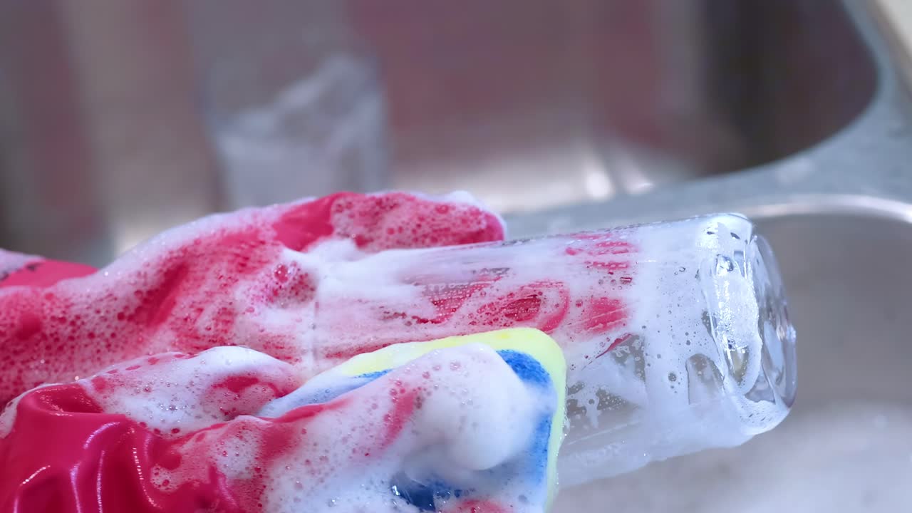 Close-up of pink-gloved hands scrubbing a glass with a soapy sponge in a kitchen sink.