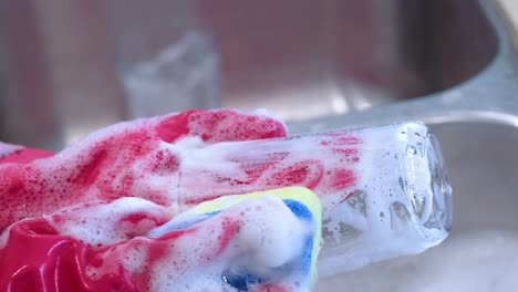 close-up of pink-gloved hands scrubbing a glass with a soapy sponge in a kitchen sink.