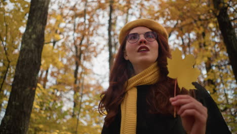 stylish lady in yellow beret twirls autumn leaf in hand while strolling through vibrant park filled with golden foliage, basking in warm sunlight and enjoying peaceful nature with graceful charm