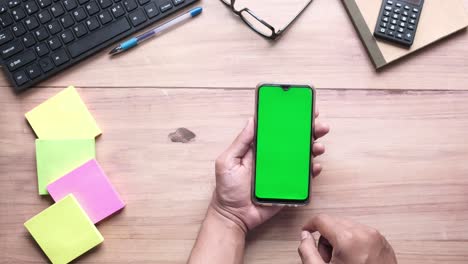 person holding a smartphone with a green screen on a wooden desk, surrounded by office supplies.