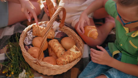 close-up of hands selecting baked snacks from a picnic basket while seated on a checkered blanket, one child drops a pastry and chooses another as sunlight highlights the warm outdoor setting