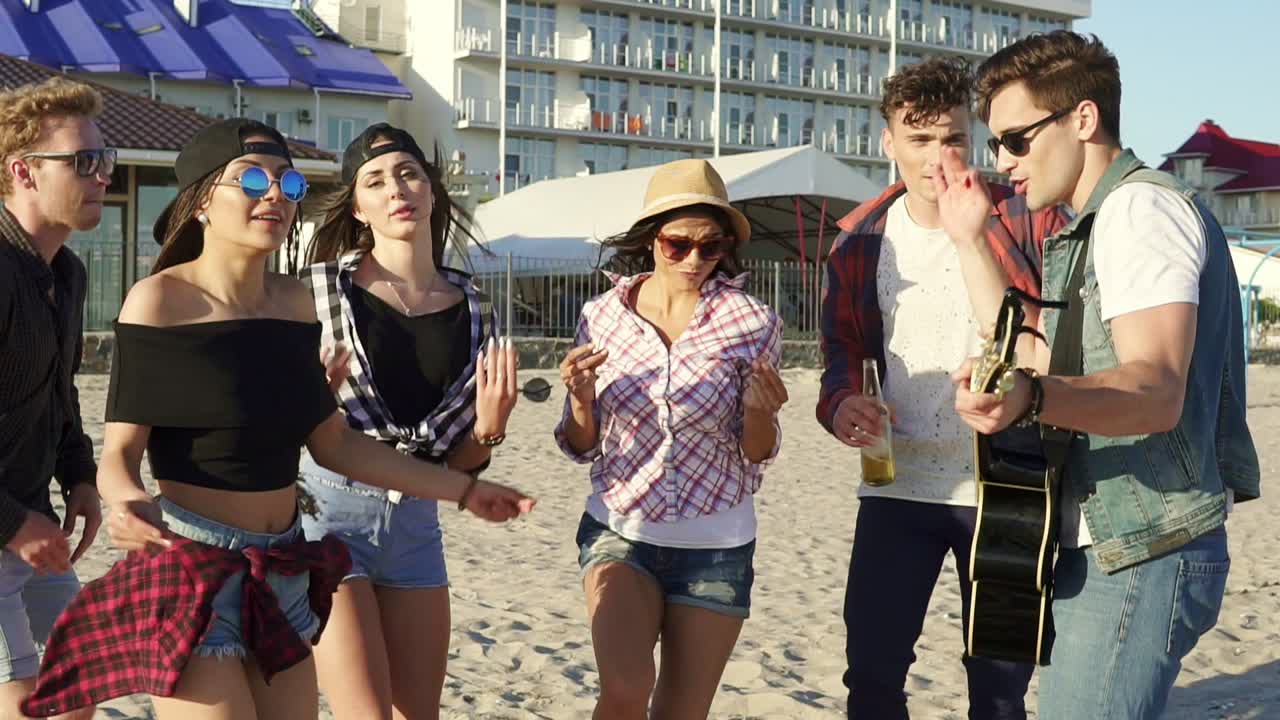 Summer party on the beach. Young boys and girls drinking cocktails, dancing together playing guitar and singing songs on a beach at the water's edge. Slowmotion shot