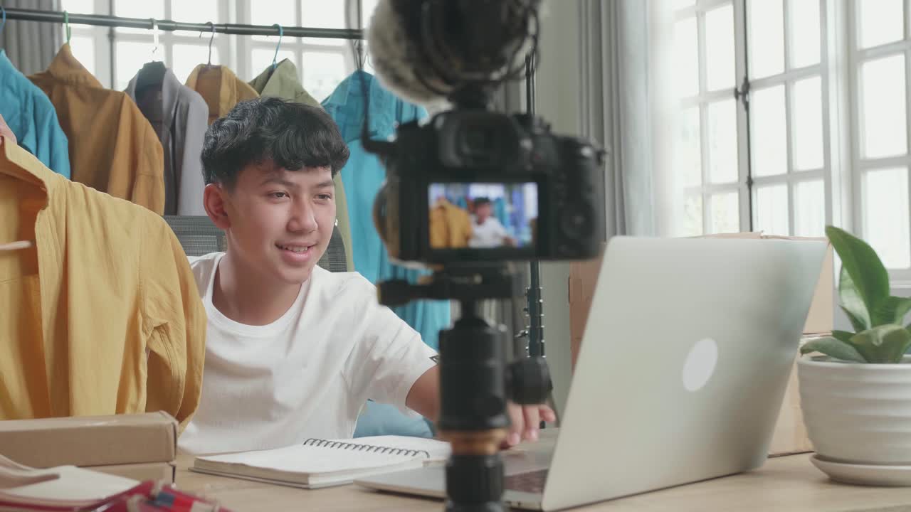 Asian Young Man Looking At Computer And Recording Video By Camera For Selling Clothes Online At Home