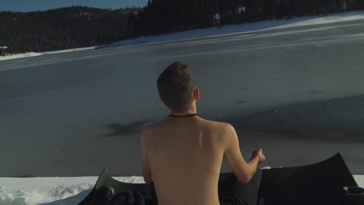 View from behind man without shirt meditating on black yoga mat on snow covered shoreline of frozen alpine lake