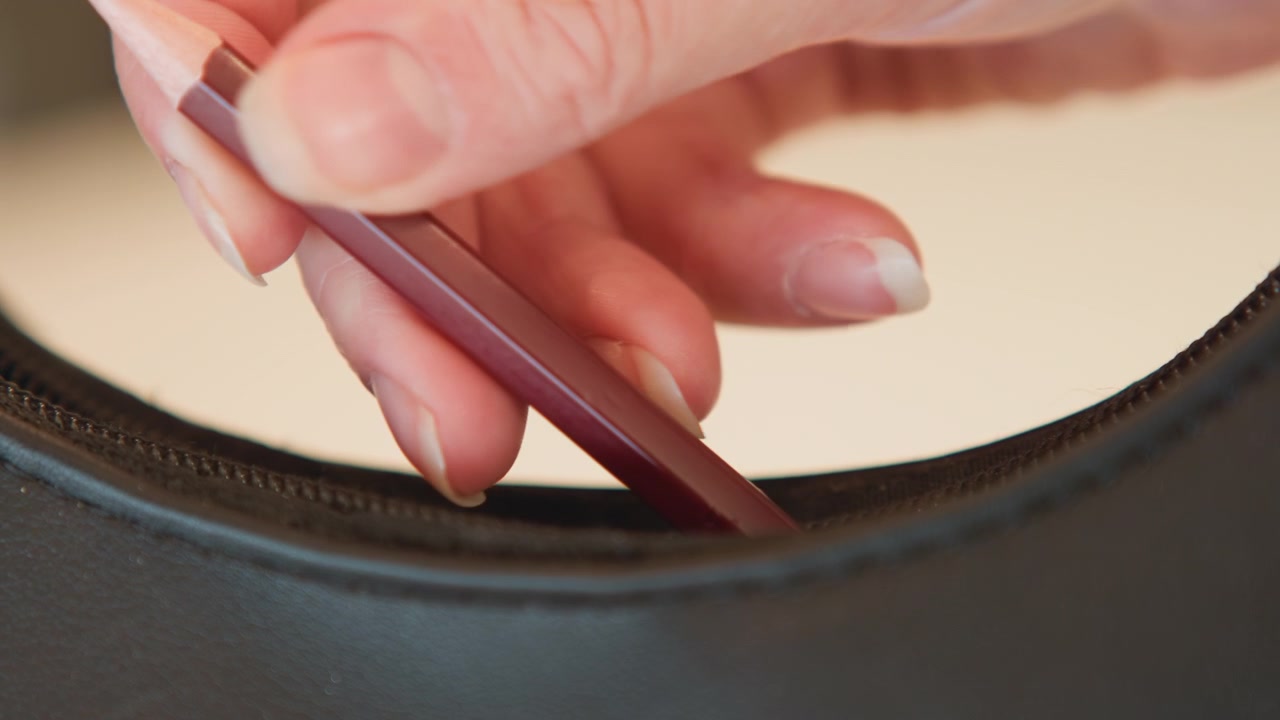 Close view shows fingertips pressing thin tool into narrow gap along curved dark leather upholstery edge, creating slight indentation, tension, and careful probing beside stitched seam