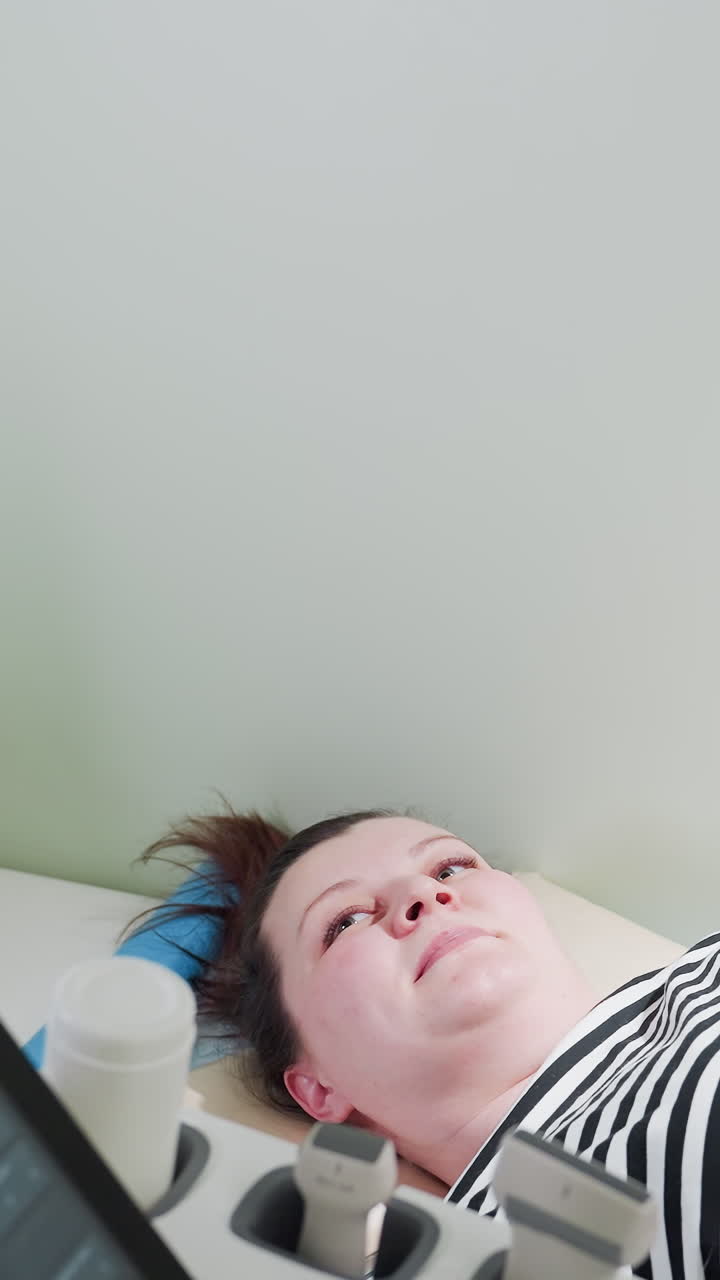 진찰대 위에 누워 있는 백인 여성, 환자는 초음파 기사가 화면을 가리킬 때 모니터를 보고 있으며, 장갑을 낀 손은 정기적인 산전 검사 중 태아 이미지를 가리키고, 초음파가 있는 은은한 조명의 병원 진료실