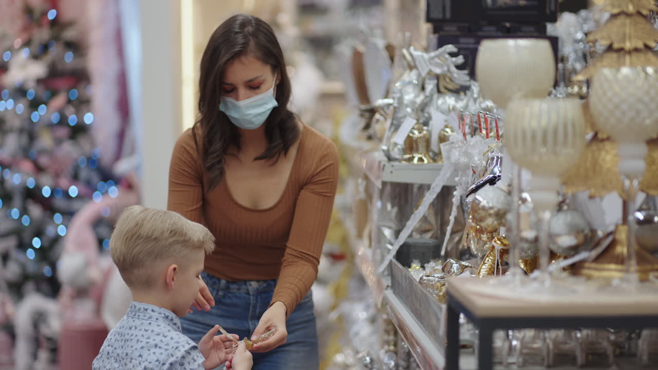 una mujer con una máscara médica con un niño en un centro comercial elige decoraciones para el hogar para navidad. mira los juguetes del árbol de navidad. epidemia de coronavirus