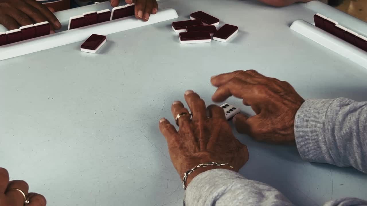Hands position two domino tiles on a light tabletop as other players hold their pieces nearby during a game in Miami