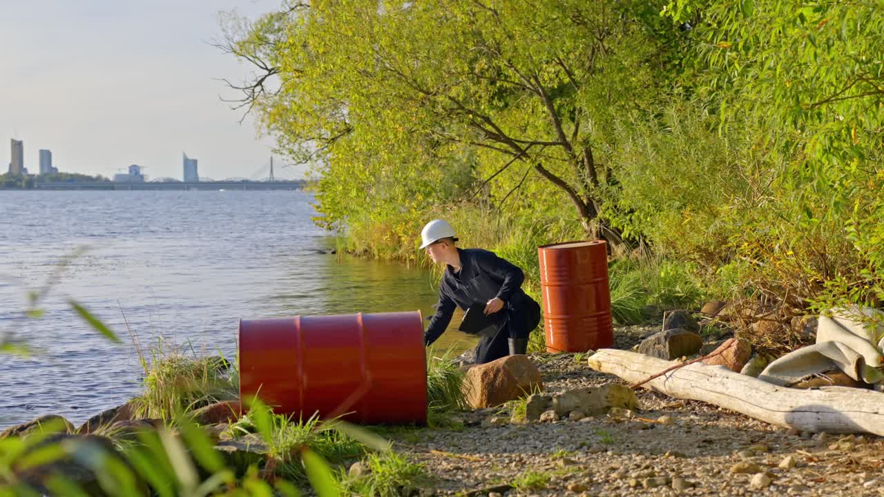 Environmental Inspector Conducting A Water Quality Test With Red Barrels On The Banks. - wide shot