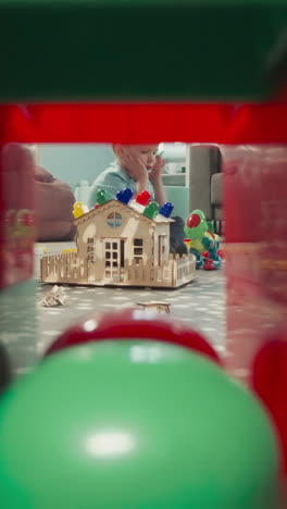 children play with toys sitting on wooden floor in nursery. girl holds baby doll while boy building house view through clearance in cube constructed wall