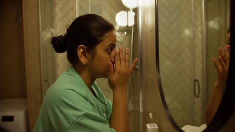 Side-view-of-a-happy-brunette-girl-in-green-pajamas-making-an-evening-mask-and-applying-special-oils-to-her-face-in-a-modern-apartment-before-going-to-bed-in-the-evening-in-the-bathroom