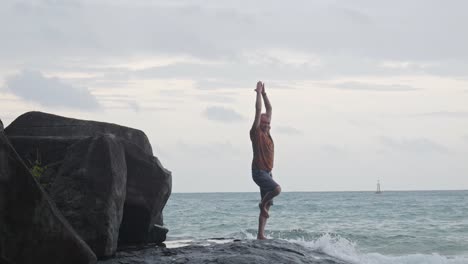 side view of man standing on one leg then make a pose in the beach with wavy ocean in the background