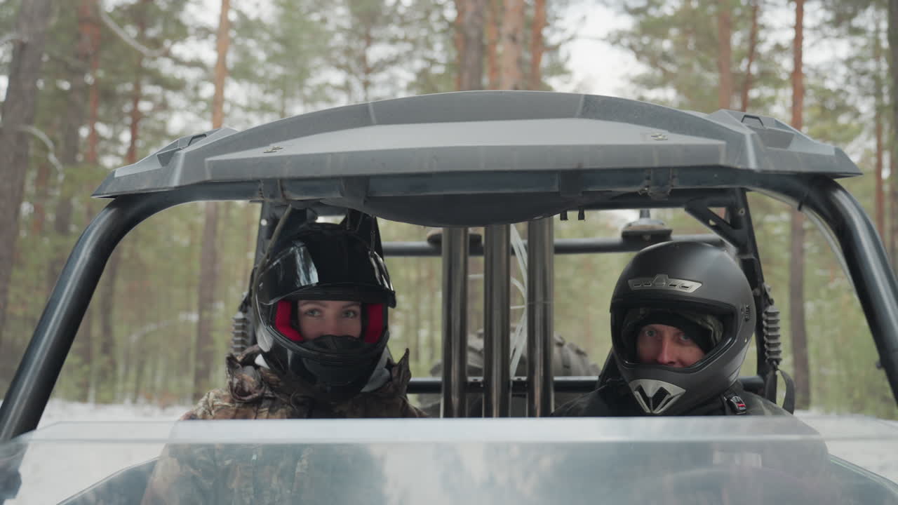 Un dúo con cascos, vistos de frente, sentados en un quad, con la mirada fija en el sendero, copilotos compartiendo anticipación y camaradería, un parabrisas despejado con escarcha, con un bosque de pinos como telón de fondo y una postura equilibrada.