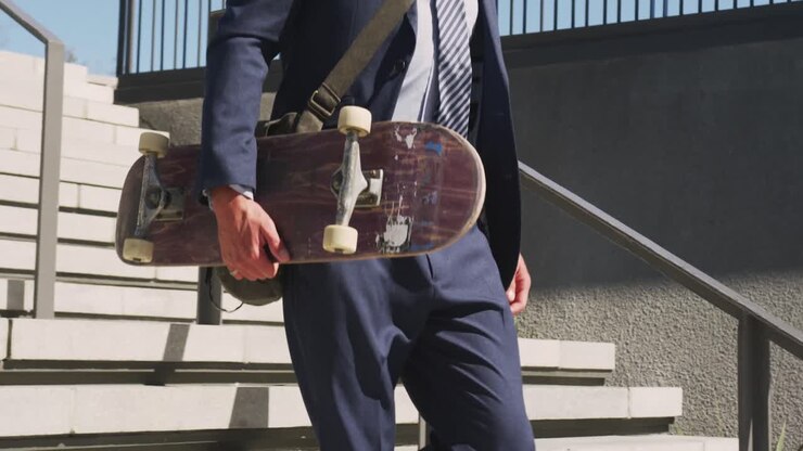 Busto di un uomo d'affari caucasico che scende le scale, con in mano uno skateboard in una giornata di sole.