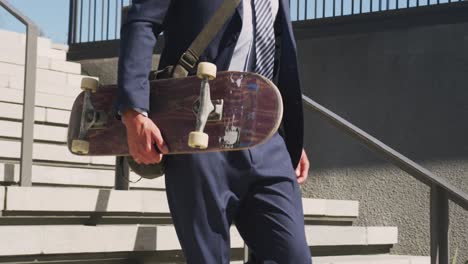 midsection of caucasian businessman walking downstairs, holding skateboard on sunny day