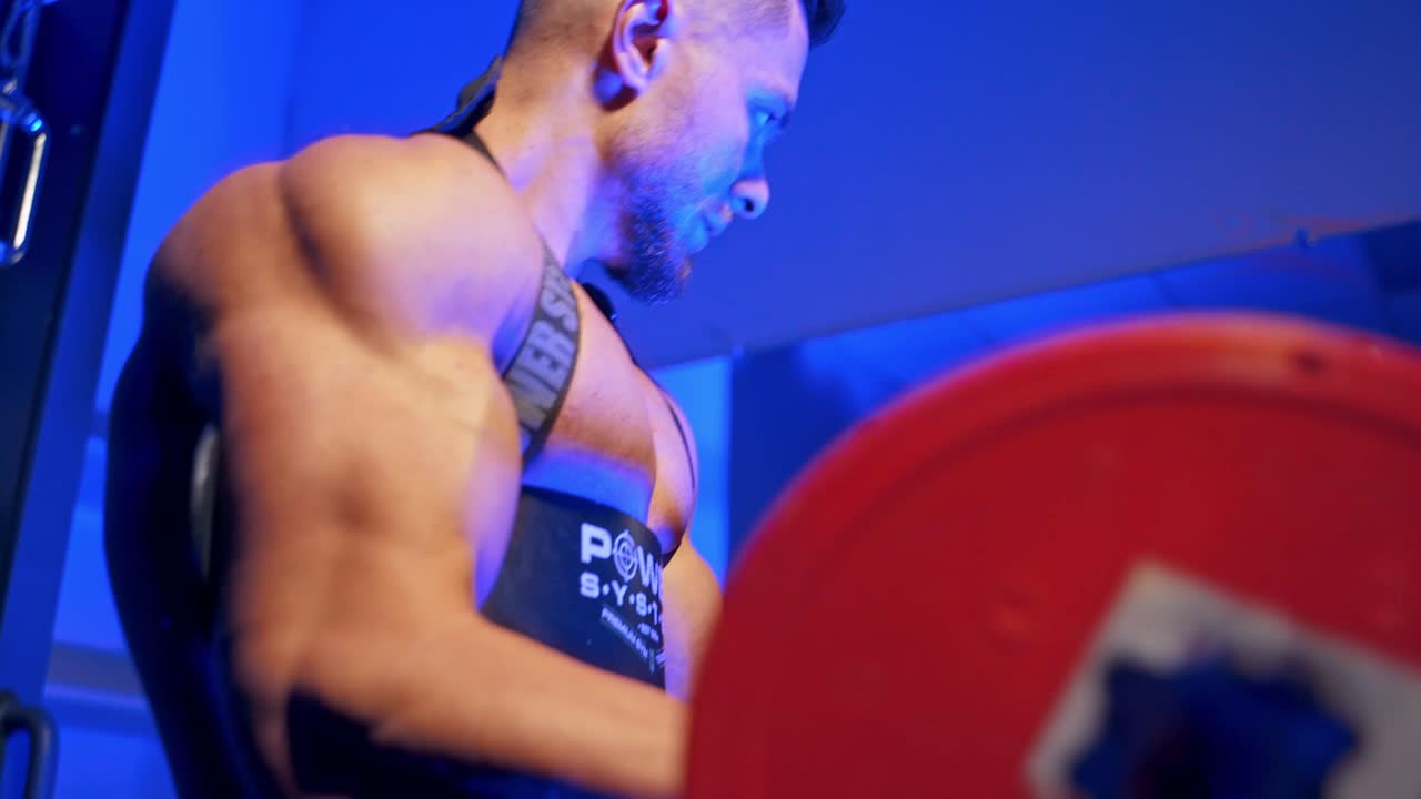 Athletic man with barbell. Muscular sportsman training his arm biceps in the gym on blue light background. Side view.