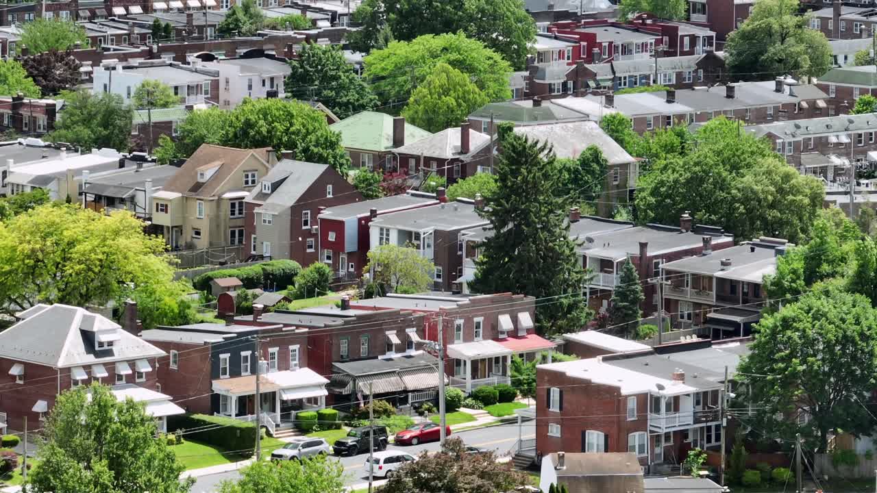 Dense American residential neighborhood with brick row houses, green trees, and backyards. Classic urban housing pattern in a quiet city area on a sunny day. Aerial wide shot.
