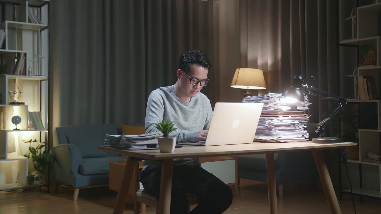 Asian Man Typing Laptop Computer While Working With Documents At Home