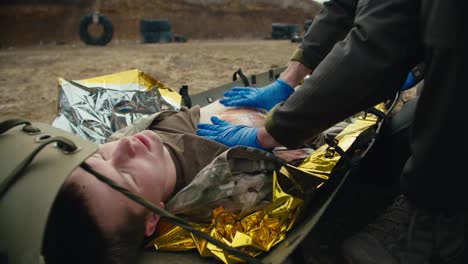 Close-up-providing-first-aid-to-wounded-soldiers-on-the-battlefield-under-the-supervision-of-a-military-medic.-Confident-male-medic-in-blue-rubber-medical-gloves-stops-the-blood-of-a-wounded-soldier-on-a-stretcher-in-a-combat-area