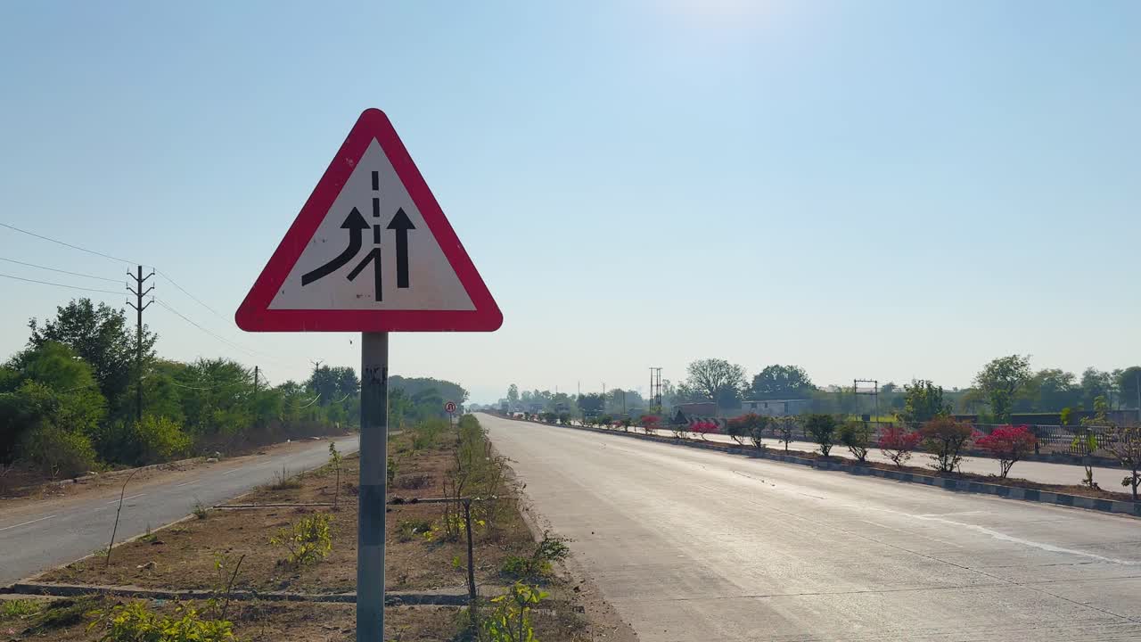 道路が分かれる高速道路の脇にある、合流する交通の警告標識、明確な車線表示、そして明るい青空の下に伸びる開けた道路を捉えた、道路脇からの静止ショット。
