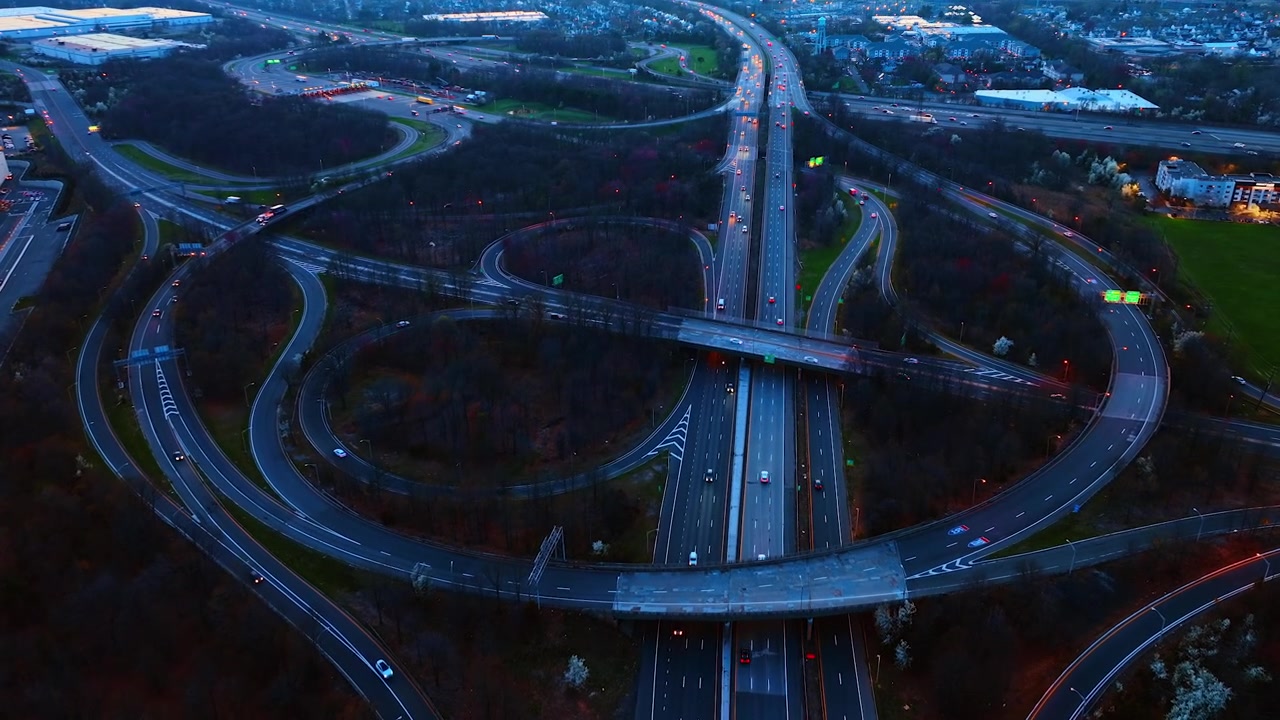Sprawling highway interchange under colorful evening sky, New Jersey. Distant elevated view of looping ramps and traffic lit at dusk with warehouses spreading toward dark hills