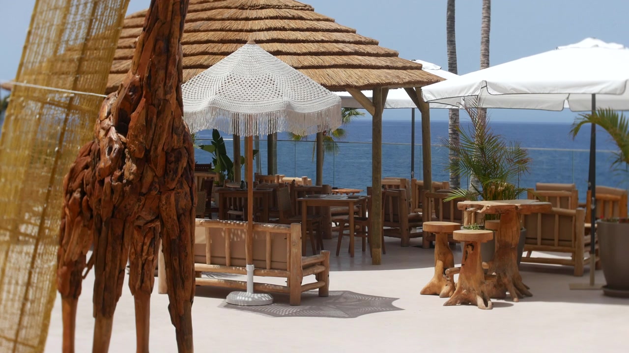 Restaurantterrasse mit Blick auf den Atlantik in Costa Adeje, Teneriffa, unter strahlender Sonne. Rustikale Holzmöbel, weiße Sonnenschirme, Palmen und Meerblick prägen die Szene.