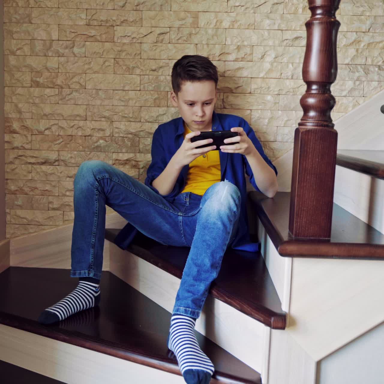 Teen boy playing on the phone on stairs. Boy sitting on a stairs at home and playing video games on his mobile phone. Gaming concept.