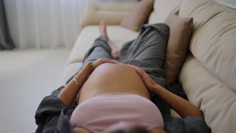 Top-view-of-happy-pregnant-brunette-woman-stroking-her-belly-while-lying-on-sofa-at-home