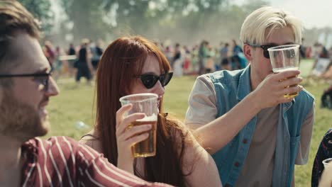 group of friends sitting on grass together at music festival and drinking beer while chatting.