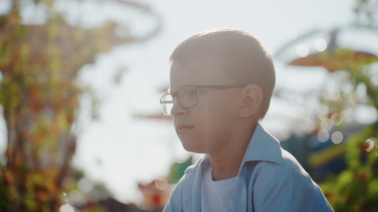 Giovane ragazzo asiatico nel parco durante una gita in famiglia nel fine settimana, con gli occhiali, che guarda in lontananza sotto il sole dorato, sfondo di fogliame delicato, umore calmo e riflessivo, illuminazione cinematografica soffusa, ritratto lifestyle