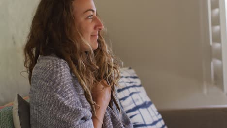 mujer caucásica sentada en el sofá con una manta alrededor de los hombros sonriendo en la sala de estar de la cabaña soleada