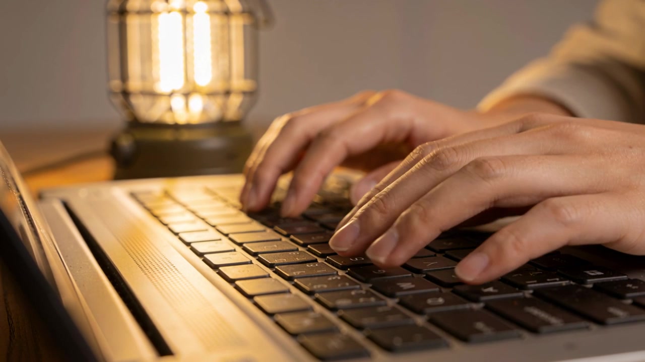 Typende volwassen handen in een lichte longsleeve die beginnen te werken, toetsen indrukkend aan een bureau thuis met een laptoplamp.