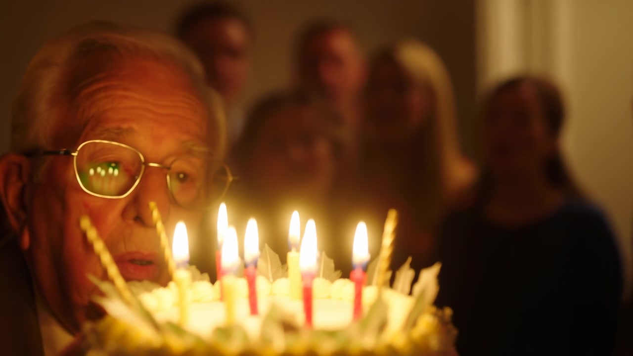 Scena in primo piano mostra un uomo anziano vicino a una torta di compleanno decorata con candele colorate accese in una luce calda e soffusa, chinato in avanti con visibile attesa durante una gioiosa celebrazione di compleanno al chiuso.