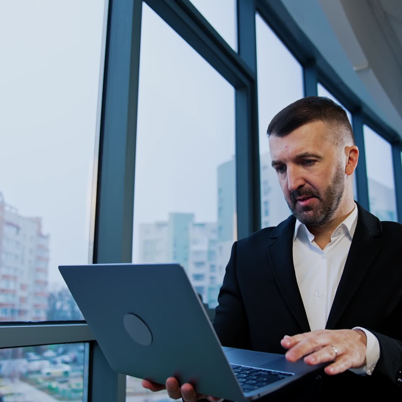Thoughtful businessman stands holding laptop in hands. Male boss types on computer waving head disapprovingly. Low angle view