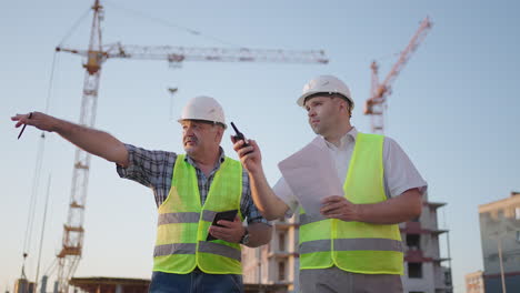 portrait of two builders standing at building site. two builders with drawings standing on the background of buildings under construction in helmets and vests