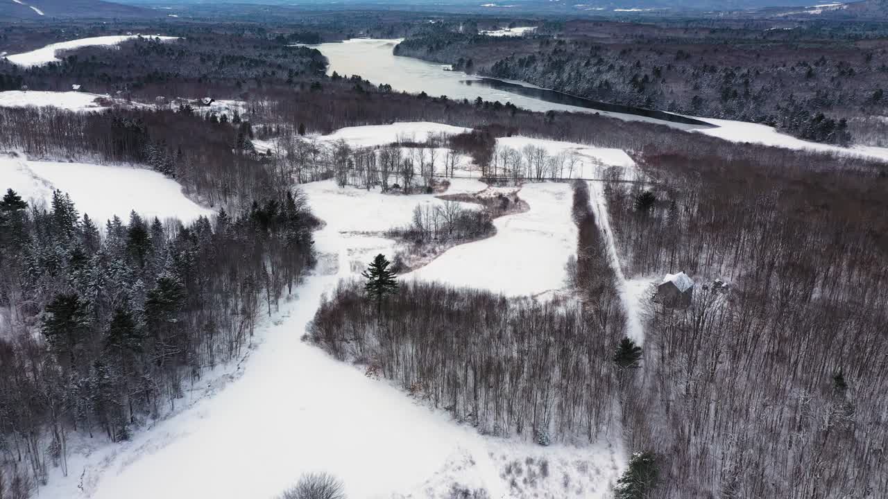 Aerial footage flying high over several snowy fields towards a partially frozen pond in a winter forest