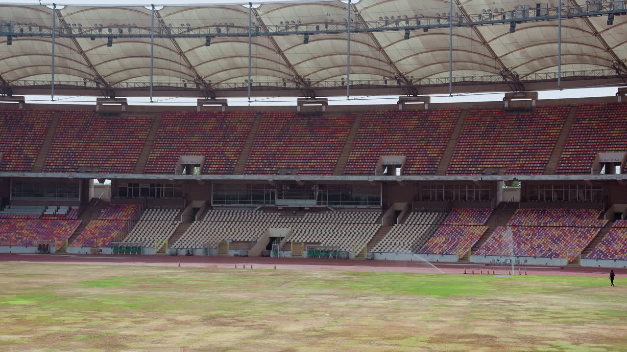 Tribune dello stadio vuote con campo in erba che mostra il manto usurato, sedili vuoti e multicolori, ampia copertura del tetto, corridoio echeggiante e tabellone segnapunti distante, calma pre-partita malinconica con cielo coperto.