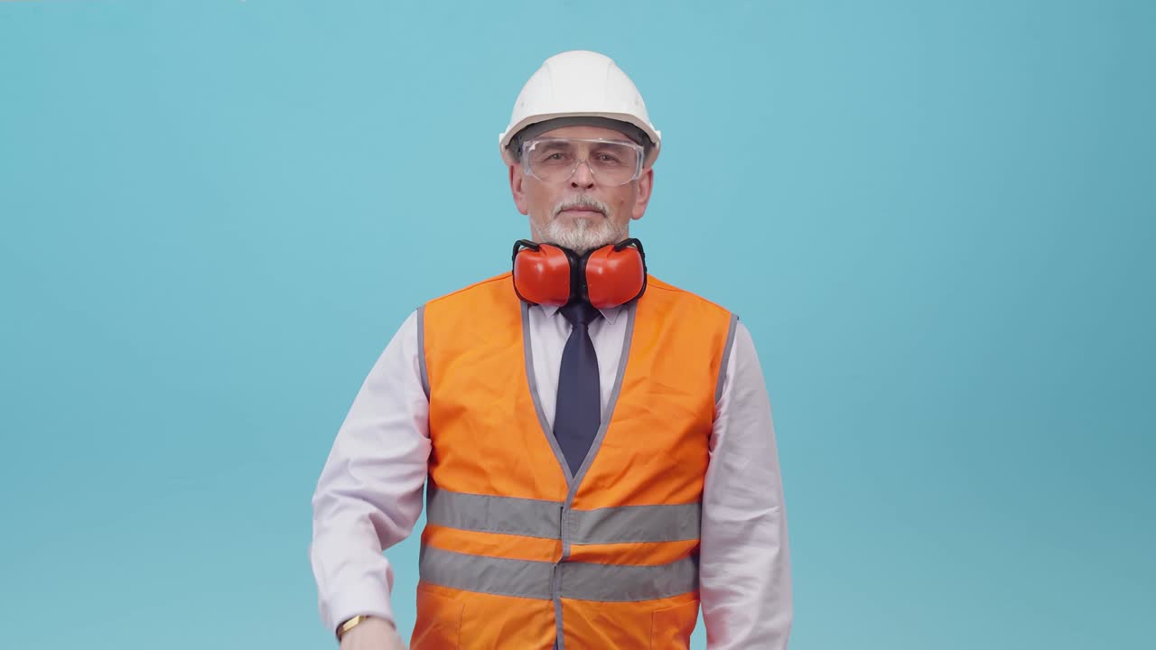 hombre adulto ingeniero con casco de protección y uniforme de trabajo muestra pantalla de teléfono con una aplicación, el fondo azul del estudio. ingeniero masculino en un chaleco de trabajo y gafas sostiene el teléfono en la mano, una pantalla verde