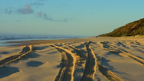 hora dorada en una playa de queensland con sombras profundas en las huellas de los neumáticos de los camiones 4x4 y un suave oleaje en el fondo