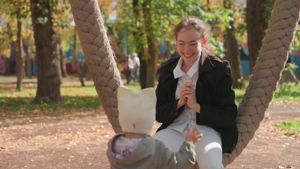 Barnepige på rebgynge tilbyder snack til lille barn i efterårspark, varm hat barn nærmer sig, latter og deling af godbid, øm omsorg og tæt familiebånd fanget i naturligt udendørs lys.