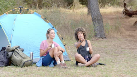 two women relax by a tent, one holding a mug and the other using binoculars