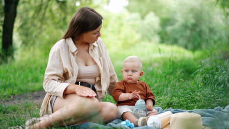 caucasian mother and little toddler son sit on the plaid outdoors. mom talks to her baby playing with a water bottle. blurred backdrop.