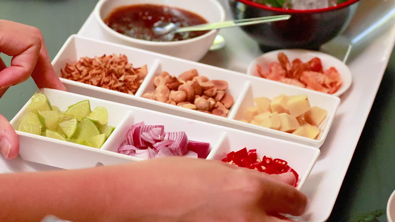Hands arrange ingredients for Miang Kham on a tray. Bright lighting highlights the vibrant colors of the traditional Thai dish