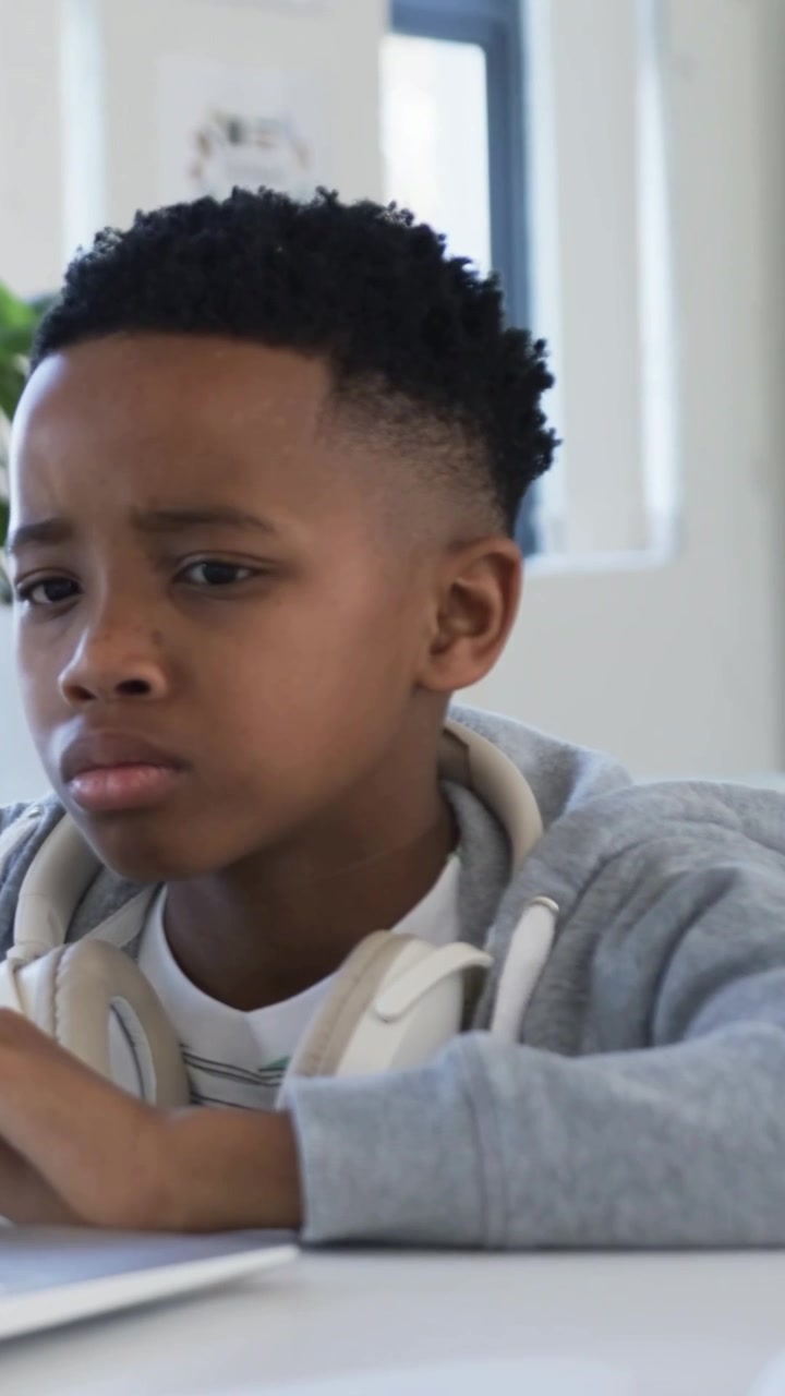 Vertical video: Leaning boy studying on laptop at home, tapping trackpad with headphones and hoodie
