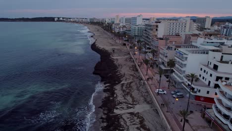 Flying-along-Cala-Millor-hotel-lined-beach-front-at-dusk,-overcast-weather