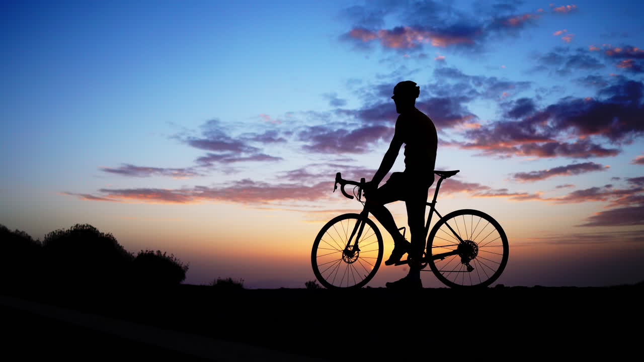 A man on a bicycle stands at the mountain's summit, enjoying the sunset as the camera smoothly glides on a Steadicam.